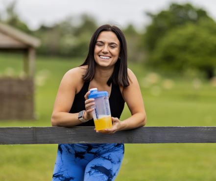 Boditronics Athlete Chelsy Osborne resting on a fence and smiling with a Boditronics shake in her hands 